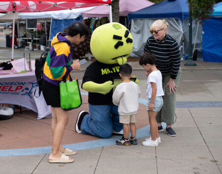 Mascot with large green head kneeling and interacting with two adults and two kids
