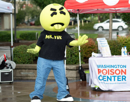 A mascot with a large green round head and a look of disgust stands in front of a red polka-dotted table canopy.