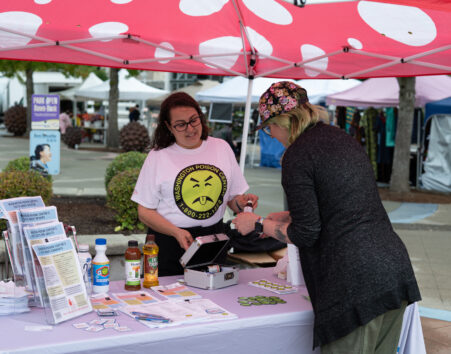 Woman behind a poison center display table, under a red and white spotted canopy talking to a another woman visting the table.