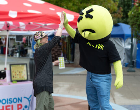 Woman in a hat high-fiving Mr. Yuk mascot, a green character with a large round head, disgusted look i=on his face, and a his tongue sticking out.