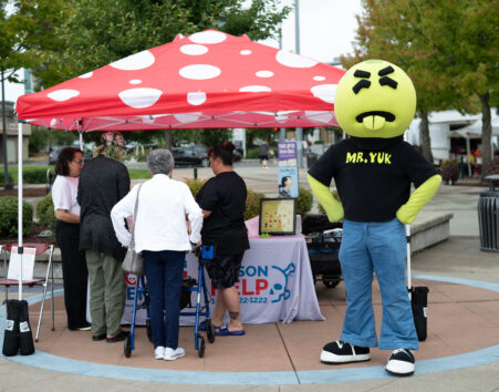 Mr. Yuk mascot, which is a green character with a big round head and a face of disgust with his tongue sticking out, standing in front of a poison center tent with a red and white spotted canopy that has several people around it with backs to the camera