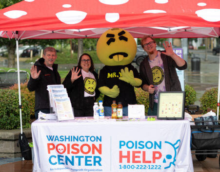 Mr. Yuk Mascot - a green character with a round head, disgusted look and sticking-out tongue), two men and a woman standing under a red and white polka-dotted tent behind a table with a poison help tablecloth waving