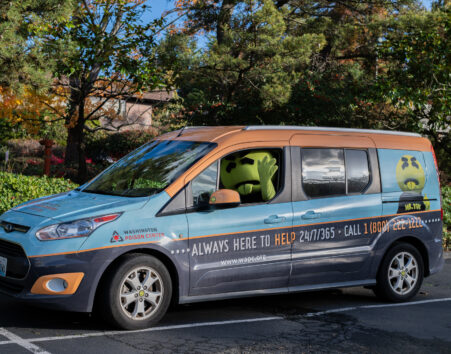 Van with a Washington Poison Center and Mr. Yuk wrap on it called the YukMobile in a parking lot with the Mr. Yuk mascot, which is a character with a large green head and his tongue sticking out, who is in the driver's seat and waving
