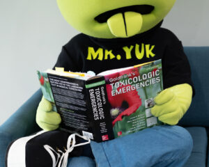 Mr. Yuk Mascot - a green character with a round head, disgusted look and sticking-out tongue) reading a toxicology book.