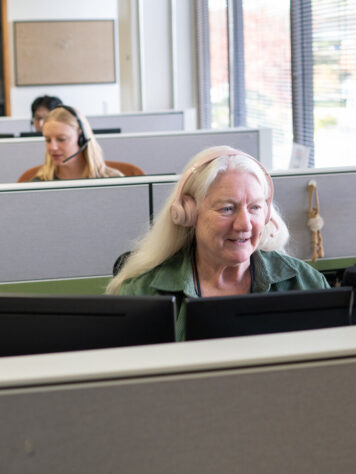 A row of people in cubicles with headsets, with an older woman smiling and talking in the foreground