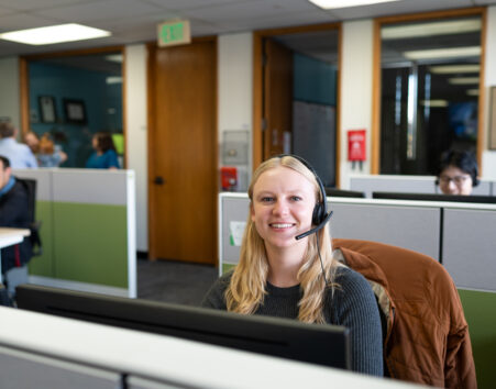 Blonde woman with a headset in a cubicle smiling at the camera