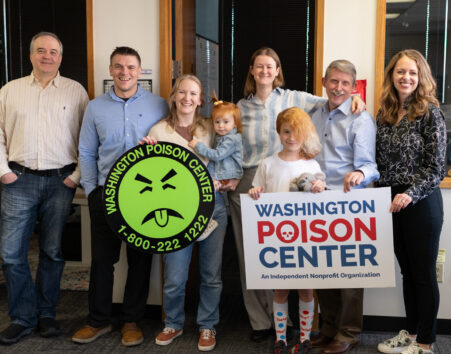 A group of 7 people of varying ages and genders holding a Washington Poison Center logo sign and a Mr. Yuk logo sign.