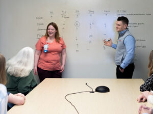 A man and a woman in nurse's scrubs at a white board presenting to a table of listeners