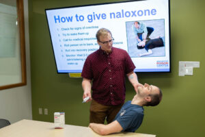 Man demonstrating how to administer naloxone in another man's nose in front of a screen that says "How to give naloxone"