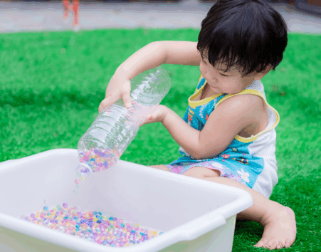Little boy pouring water beads from a bottle into a tub while sitting on a lawn.